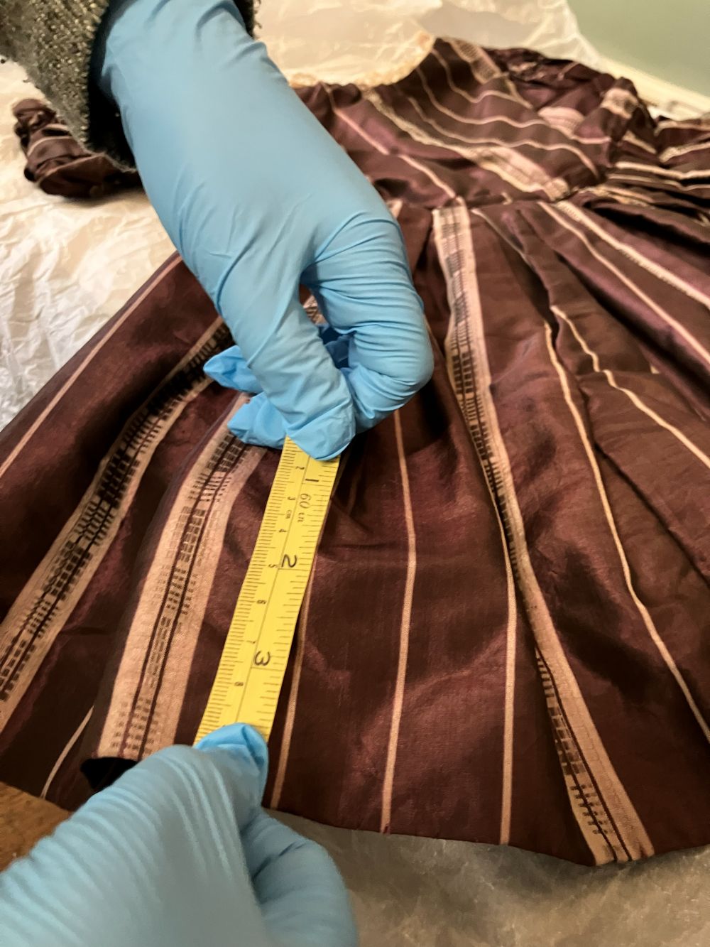 Photograph showing gloved hands using a tape measure to take measurement from red Victorian dress