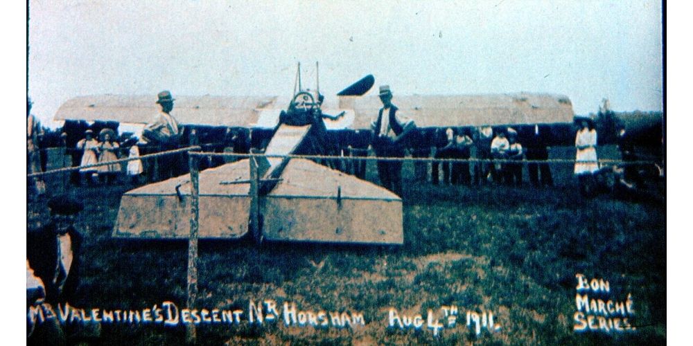 Colour postcard showing w large number of people standing under the wings of a small plane.